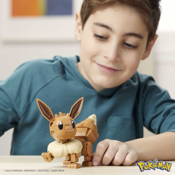 Jeune garçon souriant en sweatshirt bleu canard entrain de jouer avec la figurine en briques de construction type Lego Pokémon Evoli
